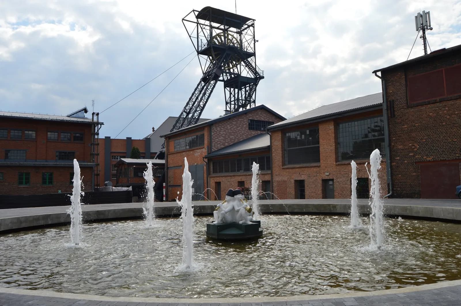 A modern fountain with jets of water spraying upwards, surrounded by historic brick buildings, with a tall dark metal mining-style tower in the background under a cloudy sky.
