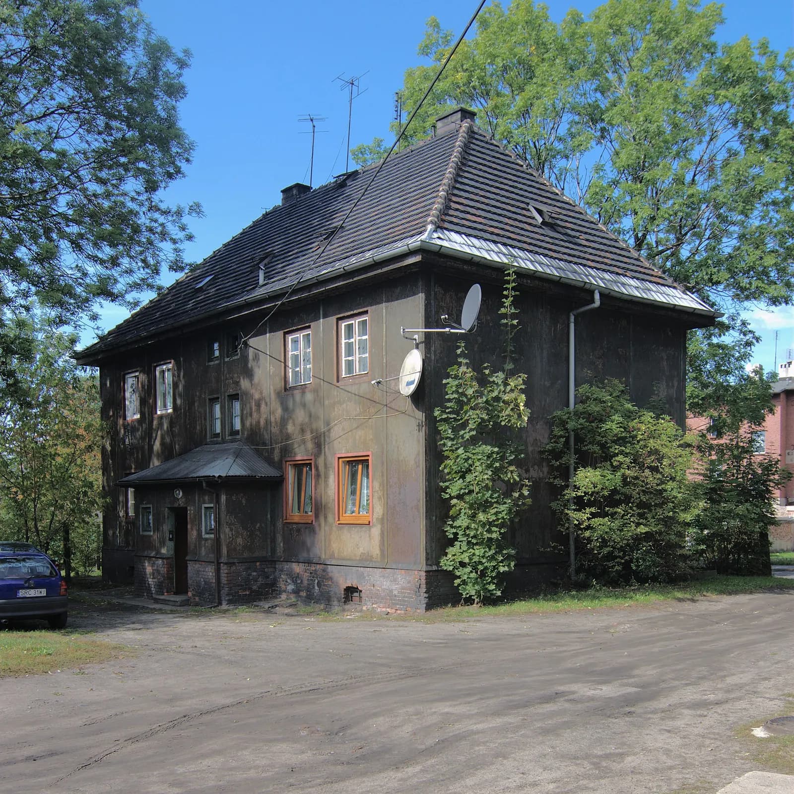 A rustic brown house with satellite dishes and a blue car parked outside, surrounded by trees under a clear blue sky.