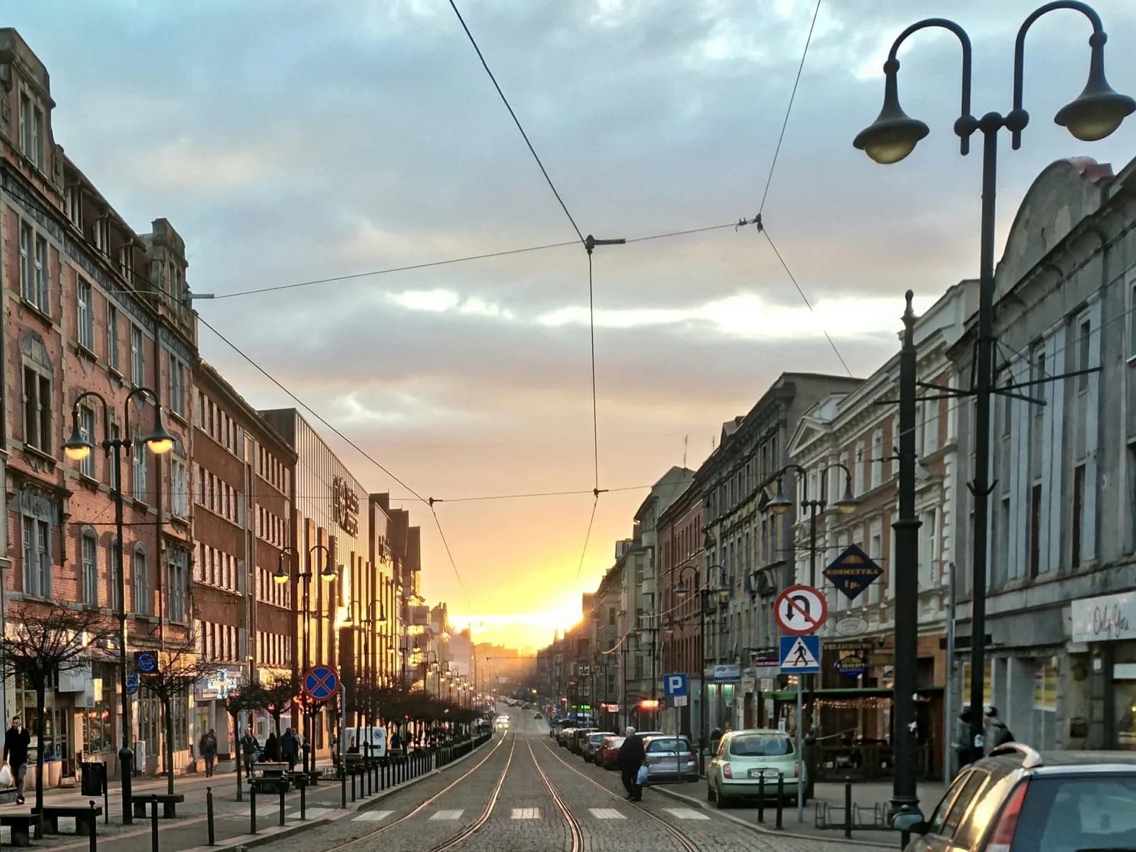 A long, straight city street with buildings lining both sides, tram tracks running down the middle, and a sunset in the distance.