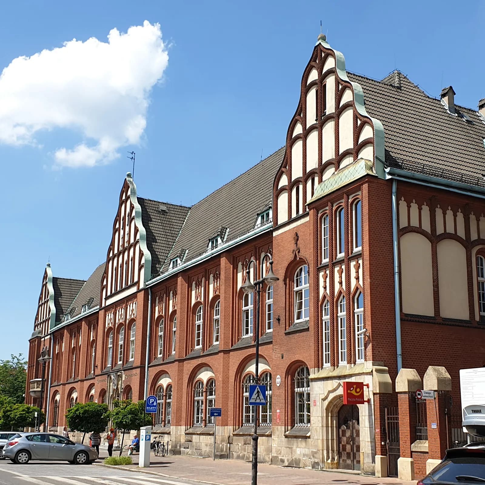 A large red brick building with multiple gables and ornate white window frames stands prominently under a bright blue sky with a single white cloud.